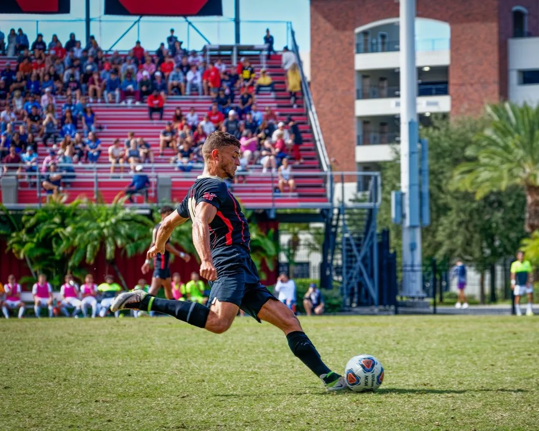 Omar Thali striking a football during a competitive college match, with a full crowd in the stands behind him