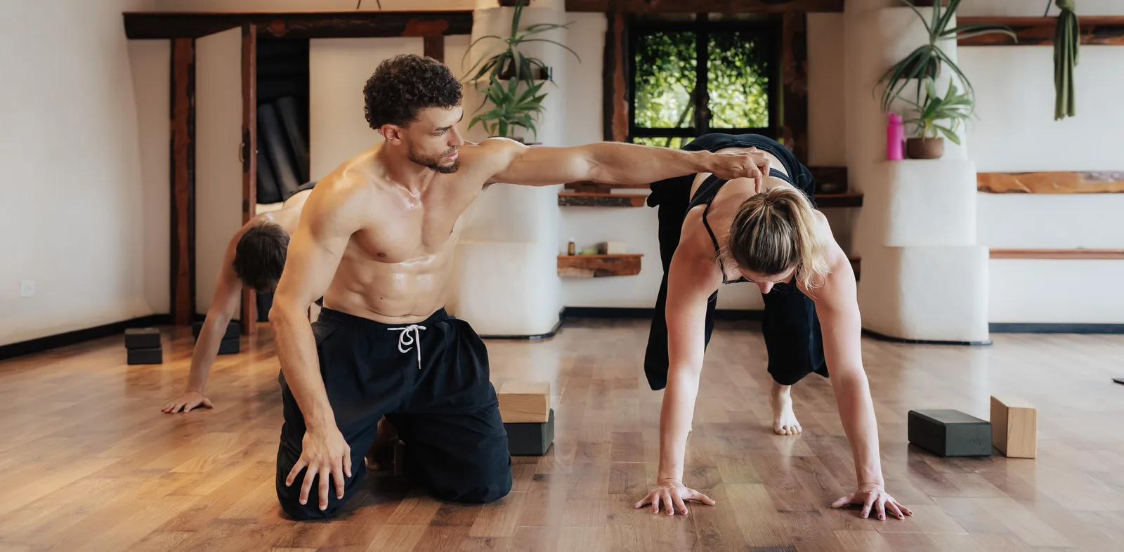 Omar kneeling beside a student in a crawl position, guiding her shoulder with his hand in a bright wooden-floored studio